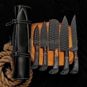 May include: A set of six black kitchen knives with wooden handles and a black leather knife roll. The blades have a textured finish and are arranged on a wooden cutting board. The knife roll is closed with a strap.