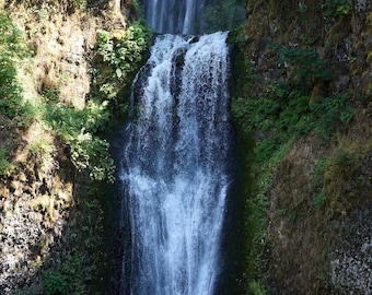 Op de afbeelding: Een waterval stort neer over met mos bedekte rotsen, wat een dramatisch tafereel creëert in een weelderig groen bos.
