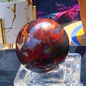 May include: A polished, spherical gemstone featuring deep red, brown, and black marbled patterns. The sphere rests on a clear, faceted glass pedestal stand, showcasing a smooth, reflective surface with natural mineral inclusions and colour variations.