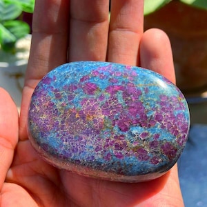 May include: A polished, oval-shaped stone featuring a vibrant mix of deep magenta-pink crystalline inclusions embedded within a smooth, mottled blue and grey matrix. The surface displays a glossy texture with varied mineral patterns throughout.