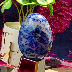 May include: A polished, egg-shaped stone featuring a mottled pattern of deep blue, white, and grey mineral inclusions. The stone is mounted on a smooth, dark, pyramid-shaped base with a metallic finish, creating a stable display for the object.