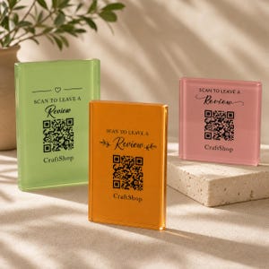 Puede incluir: Tres letreros rectangulares de acrílico en verde claro, naranja y rosa. Cada letrero presenta texto negro que dice SCAN TO LEAVE A Review, un código QR y la palabra CraftShop. Los letreros están colocados en posición vertical para mostrar la información.