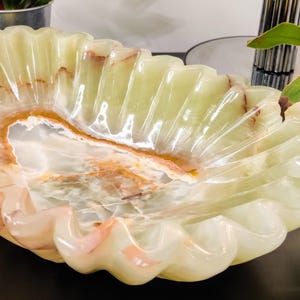 Puede incluir: Un cuenco decorativo con borde festoneado, hecho de &oacute;nix verde p&aacute;lido transl&uacute;cido. El cuenco presenta vetas naturales en tonos marrones y rosas, creando un patr&oacute;n &uacute;nico. La forma del cuenco recuerda a una concha marina.