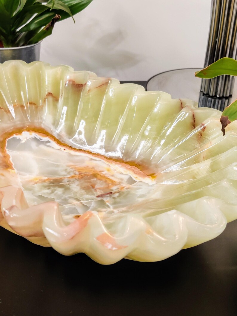 Puede incluir: Un cuenco decorativo con borde festoneado, hecho de &oacute;nix verde p&aacute;lido transl&uacute;cido. El cuenco presenta vetas naturales en tonos marrones y rosas, creando un patr&oacute;n &uacute;nico. La forma del cuenco recuerda a una concha marina.