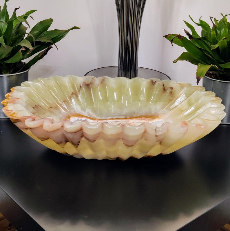Puede incluir: Un cuenco decorativo ovalado con borde festoneado. El cuenco est&aacute; hecho de un material transl&uacute;cido verde claro con vetas marrones y rosas. El cuenco se exhibe sobre una superficie negra, con plantas en macetas de metal a ambos lados.