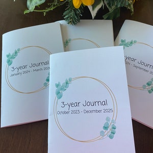 Op de afbeelding: Drie witte dagboeken met gouden en groene bloemenmotieven. De dagboeken zijn gelabeld met "3-year Journal" met de datumbereiken "January 2024 - March 2026", "October 2023 - December 2025" en "2024 - September 20".