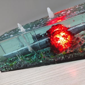 May include: A resin paperweight featuring a detailed model of a submarine submerged in water with a red explosion effect. The paperweight is illuminated from within, creating a dramatic effect.