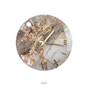 Puede incluir: Reloj de pared redondo con diseño tipo mármol, detalles dorados y números romanos. Las manecillas y los números del reloj son dorados. La esfera del reloj tiene una combinación de colores blanco, gris y dorado. La marca "RADICA" está impresa en la parte inferior.