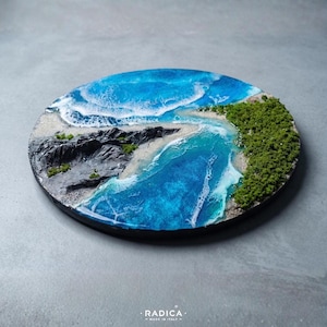 Könnte beinhalten: Rundes Kunstwerk aus Kunstharz, das eine Küstenlandschaft darstellt. Es zeigt einen leuchtend blauen Ozean mit weißen Wellen, einen Sandstrand, dunkle Felsklippen und grüne Vegetation. Ein einzigartiges Dekorationsstück.