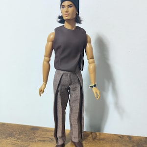 Puede incluir: Una muñeca de moda con un top sin mangas marrón oscuro, pantalones de tweed marrones y zapatos marrones oscuros. La muñeca tiene pelo largo negro, un gorro negro y una pulsera negra. La muñeca está de pie sobre una superficie de madera.