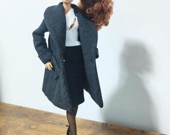 Può includere: Una bambola con un cappotto grigio scuro, una gonna e un top bianco. La bambola ha i capelli rossicci e indossa tacchi neri e calze velate. La bambola è su un supporto in acrilico trasparente.