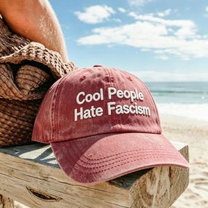 Peut inclure: Casquette de baseball rouge délavé avec l'inscription blanche "Cool People Hate Fascism". Le chapeau est posé sur une poutre en bois, avec une plage et la mer en arrière-plan.