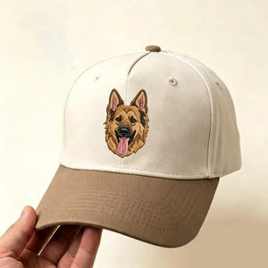 May include: A two-toned baseball cap featuring an off-white crown and a tan brim. Centered on the front panel is a detailed embroidered portrait of a German Shepherd dog with its tongue hanging out, rendered in shades of brown, black, and pink.