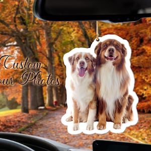 May include: A car air freshener featuring two Australian Shepherd dogs, one brown and white and the other white and brown, sitting side by side. The dogs are smiling and looking at the camera. The air freshener is hanging from the rearview mirror of a car. The background is a blurry image of autumn leaves.