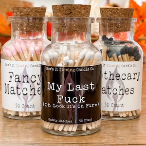 May include: Three glass jars with cork lids filled with matchsticks. The jars are labeled "Fancy Matches", "My Last Fuck (Oh Look It's On Fire)", and "Thecary Matches". Each jar has a label with the company name "How's It Glowing Candle Co."