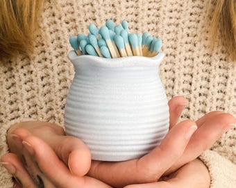 May include: A small, white ceramic match holder filled with matches. The matches have light blue tips. The holder has a ribbed design and a ruffled top. The holder is held in cupped hands.