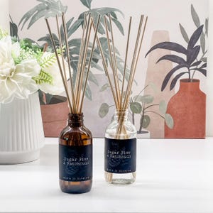 Op de afbeelding: Twee glazen diffuserflessen met riet, één amberkleurig en één helder, met het label "Sugar Pine & Patchouli". De amberkleurige fles is gevuld met een bruine vloeistof. De heldere fles is gevuld met een heldere vloeistof. De tekst "Now's It Glowing" staat eronder.