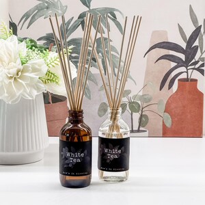 May include: Two glass diffuser bottles with reed sticks. One bottle is amber, the other is clear. Both have black labels that read "White Tea" with a floral design and the text "Now's It Glowing". The background features a floral arrangement and a botanical print.