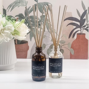 Puede incluir: Dos botellas difusoras de vidrio, una ámbar y otra transparente, con cañas. Las botellas están etiquetadas como "Grapefruit & Mangosteen". La botella ámbar está a la izquierda. El fondo presenta un estampado floral y un jarrón blanco con flores blancas.