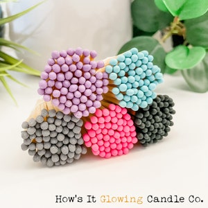 May include: A close-up of a bundle of colorful matches with round, textured heads. The colors are purple, blue, pink, gray, and black. The matches are arranged in a semi-circle with the text "How's It Glowing Candle Co." at the bottom of the image.