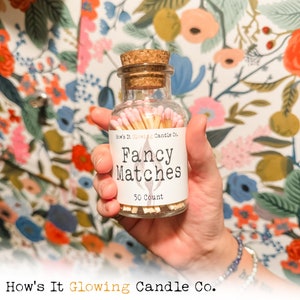 May include: A glass jar filled with 50 pink and yellow matches. The jar has a cork lid and a white label that reads "How's It Glowing Candle Co. Fancy Matches 50 Count".