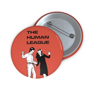 May include: Two red, circular badges with pin backs. One badge displays the text "THE HUMAN LEAGUE" above a black and white illustration of two dancing figures. The other badge is silver.