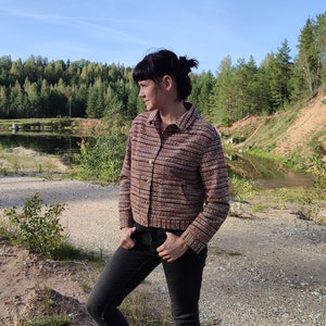 May include: A woman wearing a multi-colored striped jacket with black jeans and black ankle boots. She is standing in a gravel area with trees and a pond in the background.