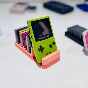 Può includere: Una console di gioco portatile Game Boy Color verde lime con una selezione di cartucce di gioco in un supporto color corallo. La console ha pulsanti neri e il testo "GAME BOY COLOR" sullo schermo.