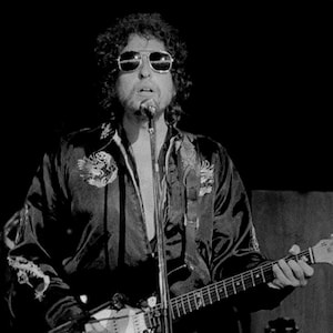 May include: A black and white photo of a musician wearing sunglasses and a jacket, playing a white electric guitar with a microphone in front of him.