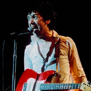 Puede incluir: Un hombre con el pelo oscuro y una camisa blanca canta en un micrófono mientras sostiene una guitarra eléctrica roja y blanca.