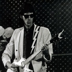 May include: A black and white photo of a musician playing a white electric guitar with a lightning bolt design on the front. The musician is wearing a white blazer with a black shirt and a black tie. The musician is wearing sunglasses.