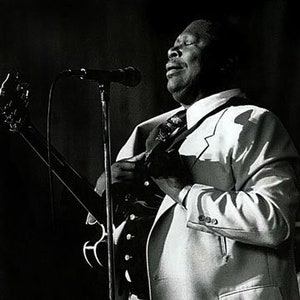 May include: A black and white photograph of a man in a white suit playing a guitar and singing into a microphone.