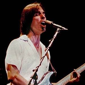 May include: A man with long brown hair sings into a microphone while playing a silver electric guitar. He is wearing a white shirt and jeans.