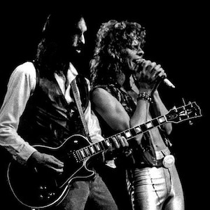 Op de afbeelding: Twee muzikanten die op het podium optreden, de een speelt een zwarte elektrische gitaar en de ander zingt in een microfoon.