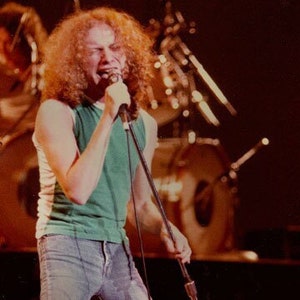 Puede incluir: Un hombre con el pelo largo y rizado canta en un micrófono mientras lleva una camiseta sin mangas verde y pantalones vaqueros azules. Está actuando en un escenario frente a una multitud.