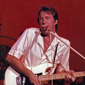 Peut inclure: Un homme en chemise blanche joue de la guitare électrique blanche avec un pickguard noir. Il porte un pantalon beige et a un microphone devant lui.