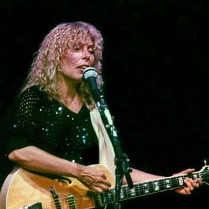 Pode incluir: Uma mulher com cabelo loiro encaracolado canta em um microfone enquanto toca uma guitarra elétrica de corpo oco marrom e preto.