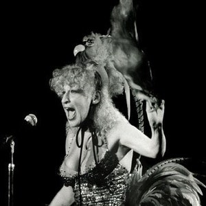 May include: A woman wearing a sequined dress and a feathered headdress performs on stage with a microphone.