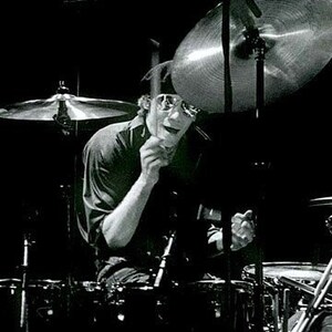 Może przedstawiać: Czarno-białe zdjęcie perkusisty grającego na perkusji. Perkusista ma na sobie okulary przeciwsłoneczne i ciemną koszulę. Widoczne są talerze i zestaw perkusyjny. Zdjęcie zrobione z niskiego kąta, z perkusistą w centrum.