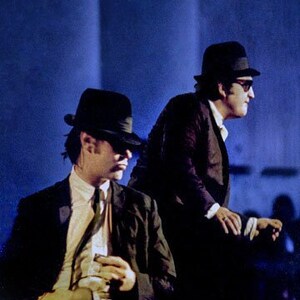 May include: Two men wearing black suits and black fedora hats. The man in the foreground is holding a microphone in his right hand.