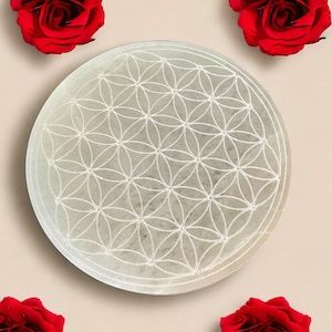 Puede incluir: Un plato de carga de selenita blanco, redondo, con un diseño de flor de la vida tallado. El plato está rodeado de rosas rojas.