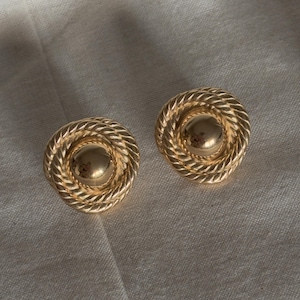 May include: A pair of gold-coloured clip-on earrings. Each earring has a central, rounded stud encircled by a rope-like, spiral design. The earrings are set against a neutral, textured background.