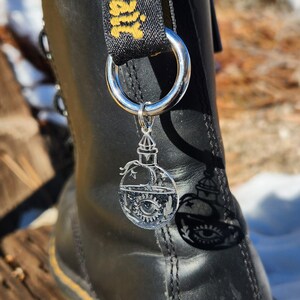 May include: Black leather boot with a yellow and black strap and a silver charm attached to a silver ring. The charm is a silver bottle with a moon and stars design and an eye in the center.