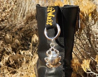 May include: Black leather boot with a yellow and black Dr. Martens logo on the side. A silver charm with a beetle design is attached to the boot with a silver chain.
