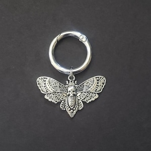 May include: Silver-toned keychain featuring a detailed moth pendant. The moth design incorporates a skull motif on its body and intricate wing patterns. The keychain has a round clasp for easy attachment, set against a dark background.