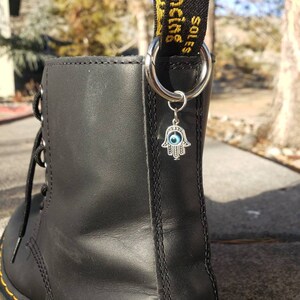 May include: Black leather boot with a yellow stitch detail and a silver charm attached to the laces. The charm is a hamsa hand with a blue evil eye in the center. The laces have a black and yellow ribbon attached with the text "SOLES WITH BOUNCING LIFE" printed on it.