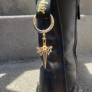 May include: Black leather boot with a gold chain and a gold dagger charm. The charm has a star and a circle design. The boot has a yellow stitching detail around the sole.