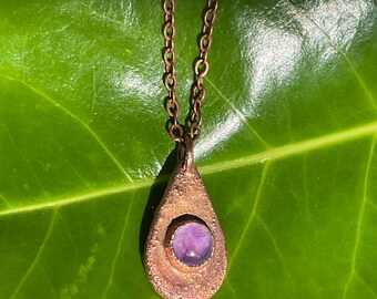 May include: A copper teardrop pendant necklace with a purple amethyst gemstone set in the center. The necklace is photographed on a green leaf.