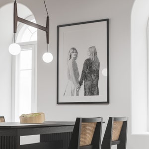 May include: A black framed drawing of two women standing side by side, holding hands. The drawing is in a minimalist style, with a white background and black lines.