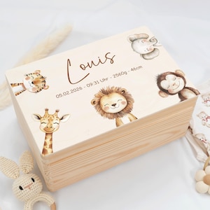 Pode incluir: Uma caixa de lembranças de madeira clara com uma tampa decorada com animais de desenho animado, incluindo um leão, uma girafa e um macaco. A tampa tem o nome "Louis" e detalhes do nascimento. Uma chocalho de coelho de crochê e roupas de bebê estão por perto.
